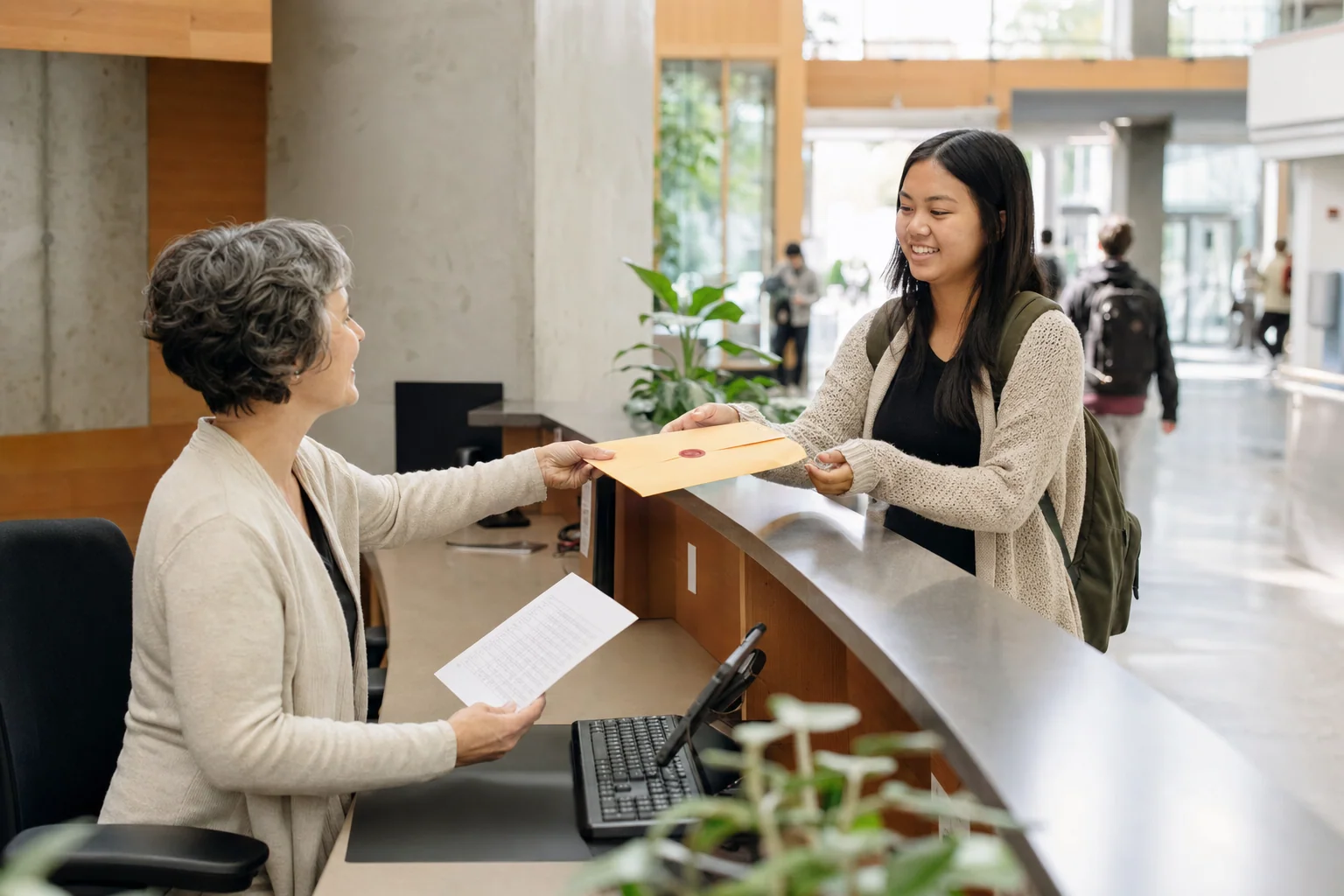 At a university registrar's desk, a staff member hands a student a sealed certificate envelope while checking a printed record.