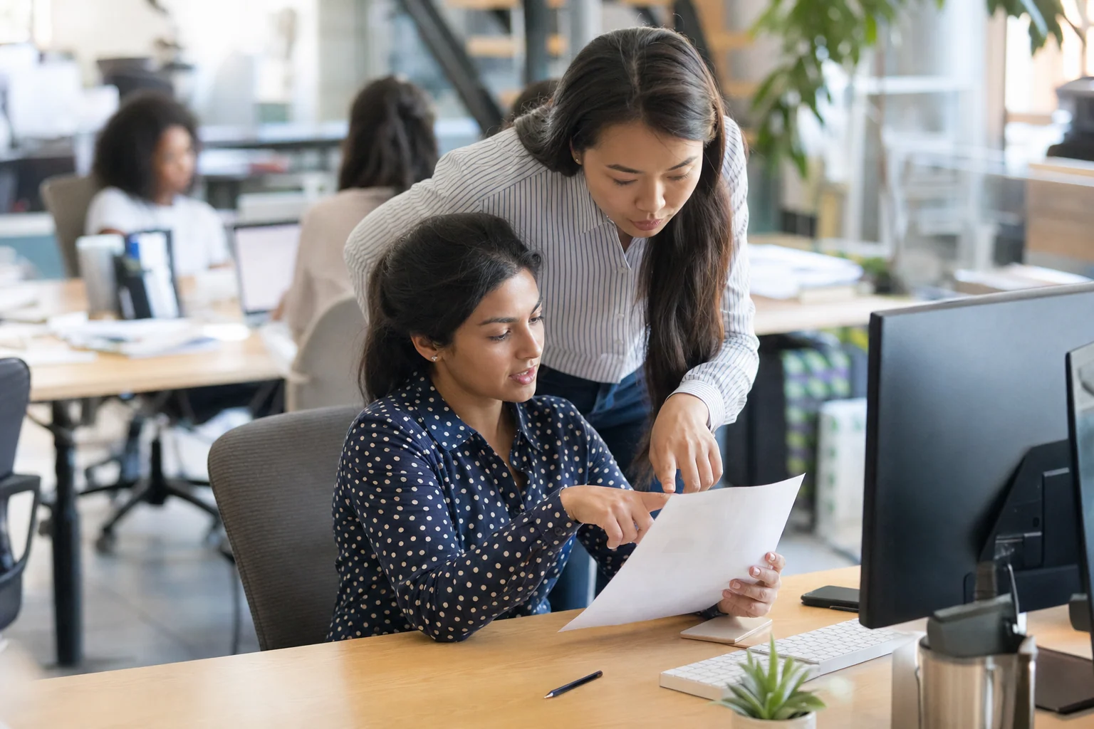An accounts payable specialist reviews a printed invoice with a colleague from purchasing who points at a line on the page in a bright office.