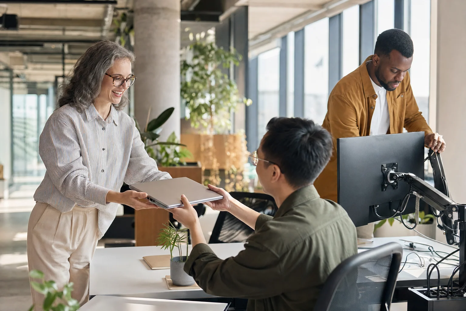 An HR partner welcomes a new employee and hands over a laptop while an IT colleague sets up the desk in a bright office.