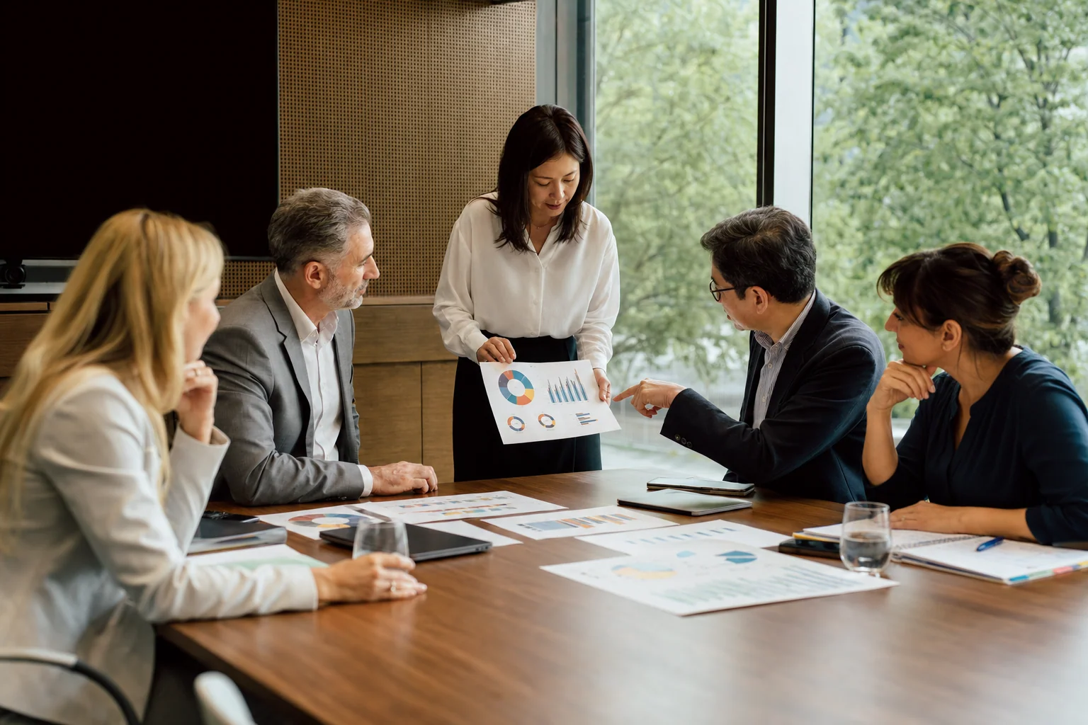 In a monthly management review, a branch manager presents printed charts to a leadership team around a boardroom table.