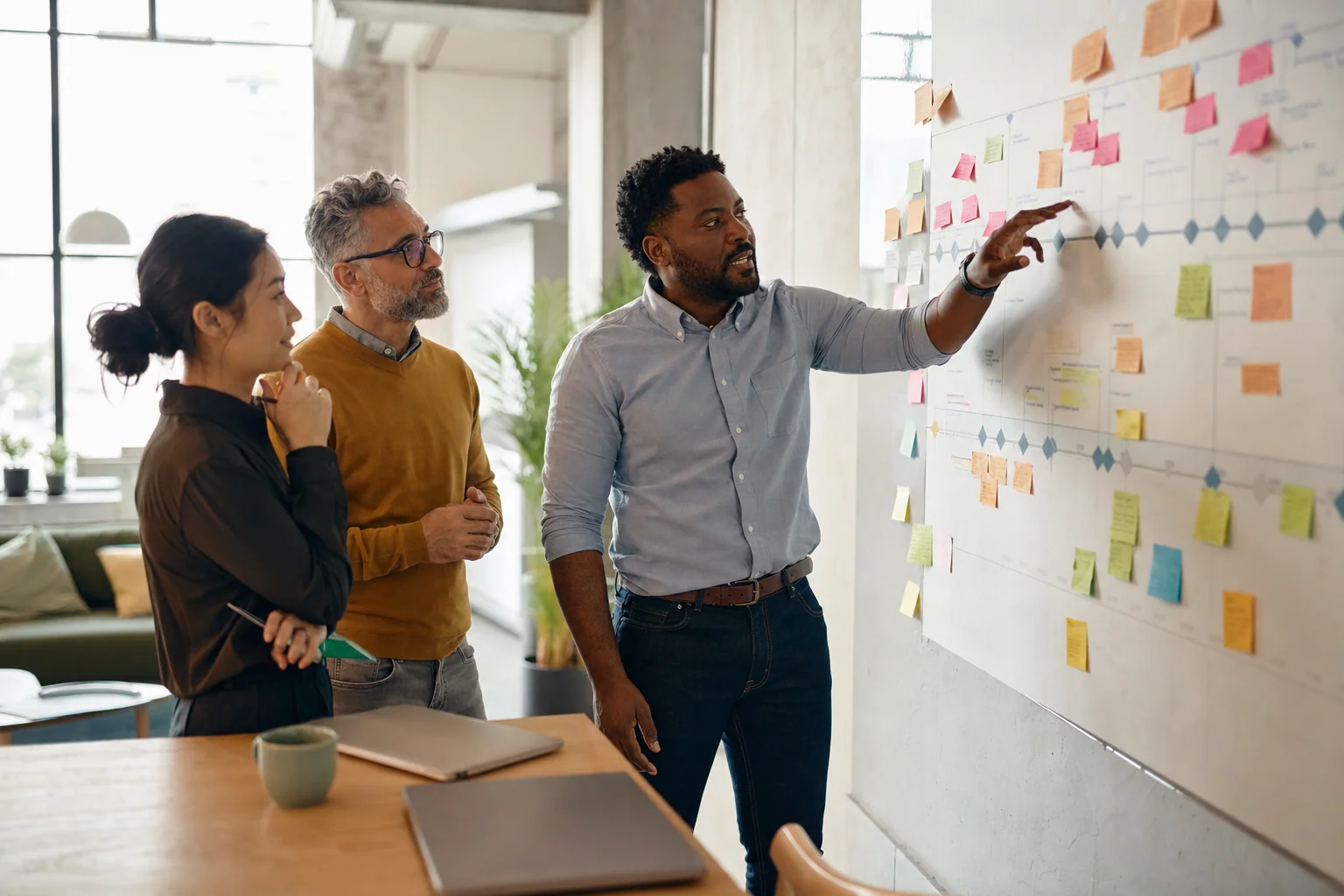A project lead points at a milestone on a wall plan covered with sticky notes while two teammates discuss it in a bright studio.